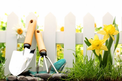 A close-up of a section of a garden in Stoke Newington, featuring a small patch of vibrant green grass bordered by a white picket fence in the background. In the foreground, there are yellow daffodils with green leaves growing beside the grass. To the left, two gardening tools—a hand trowel and a pair of pruning shears—rest against the soil, which appears freshly prepared for planting. The scene is illuminated by natural daylight, suggesting a clear weather day, and the overall setting emphasizes well-maintained outdoor space suitable for gardening and landscaping services provided by Gardener Stoke Newington. This garden area shows a combination of natural plant growth and prepared soil, indicative of ongoing gardening work aimed at enhancing residential outdoor environments in the Stoke Newington area.