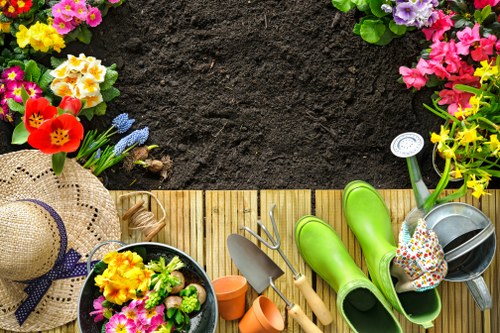 A view of a section of a garden at the edge of a wooden deck, featuring various gardening tools and accessories. The foreground includes bright yellow, pink, and purple flowering plants in pots, with a straw sun hat placed nearby. On the deck, there are green rubber gardening boots, a metal watering can with a colorful polka-dotted fabric cover, small terracotta plant pots, and hand gardening tools such as a trowel and fork. The background shows freshly turned dark soil, bordered by a row of flowering plants and bushes with green foliage. The scene appears to be a well-maintained outdoor space, suitable for gardening activities, in a residential area consistent with Stoke Newington, London, highlighting a typical garden setup ready for planting and outdoor care. The weather looks clear and bright, suggesting a sunny day ideal for gardening work, exemplifying the kind of outdoor environment that Gardener Stoke Newington services can help maintain and improve.